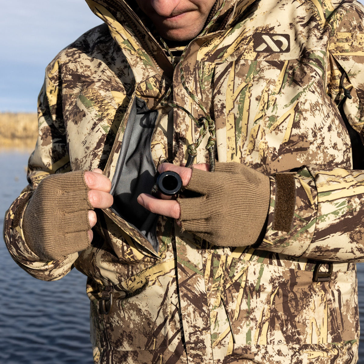 Close-up of a person wearing a First Lite marsh camouflage hunting jacket and tan fingerless gloves, holding a black duck call with the jacket's zipper open.
