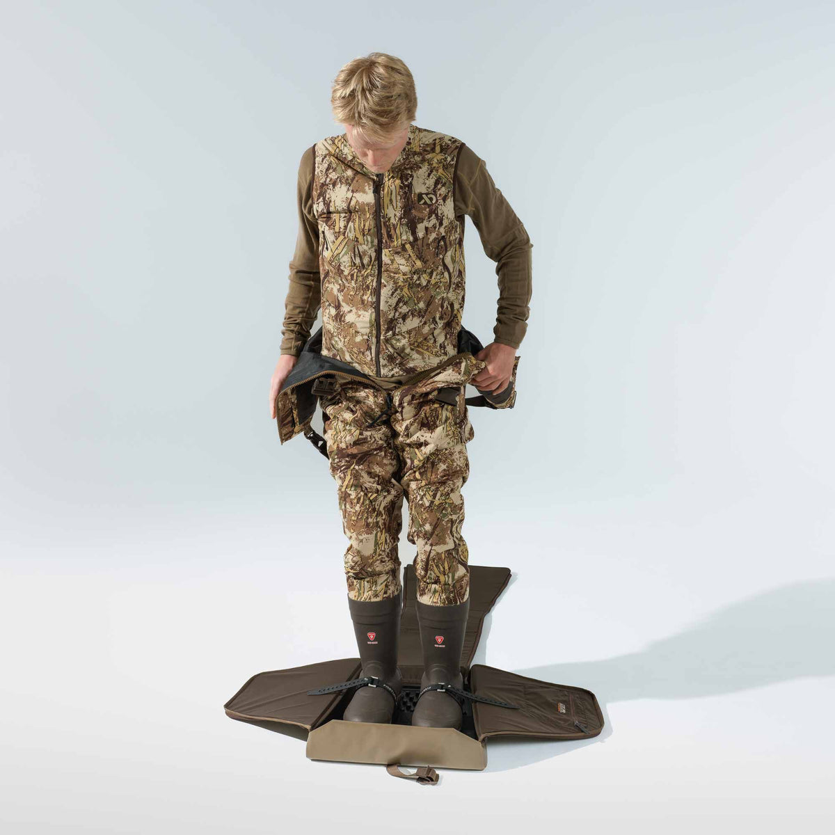 A person wearing a camouflage hunting bib and vest with dark brown boots stands on a brown portable boot-changing mat, unfastening their bib.