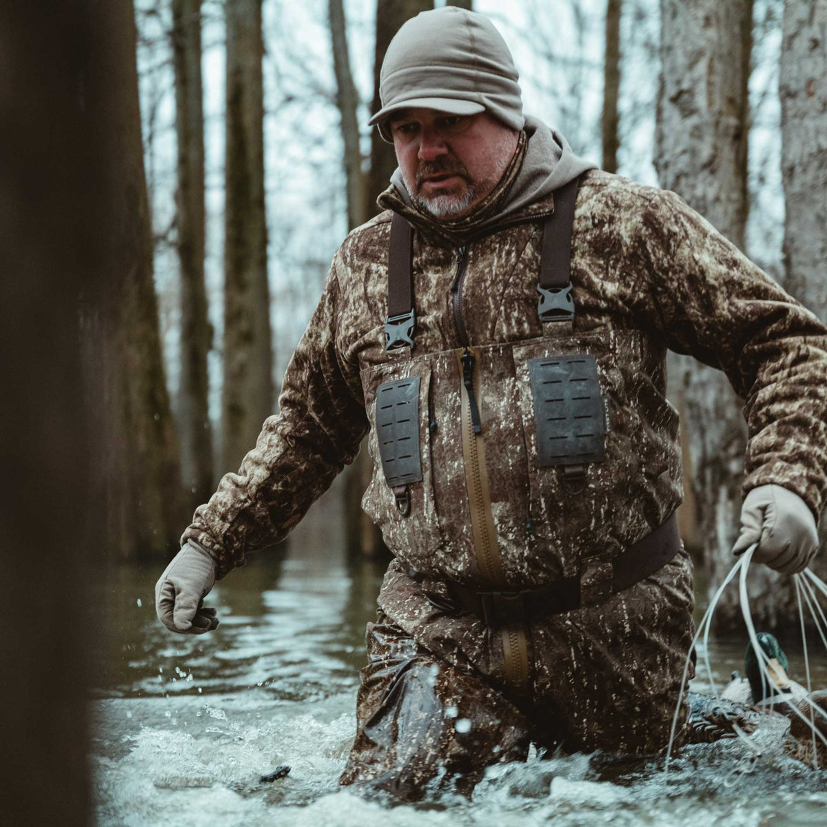 Brown camouflage wader suit with suspenders, a front zipper, and chest utility panels, shown on a man walking through water.