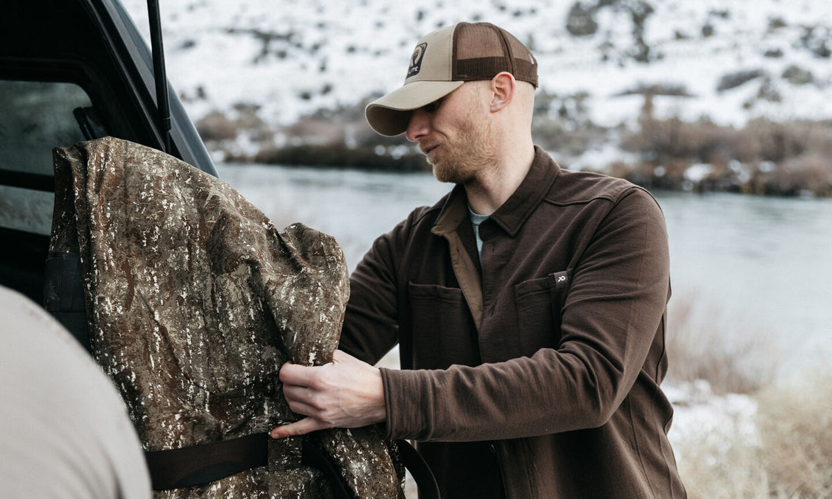 Man pulls a brown speckled camouflage blind from a vehicle by a snowy river.