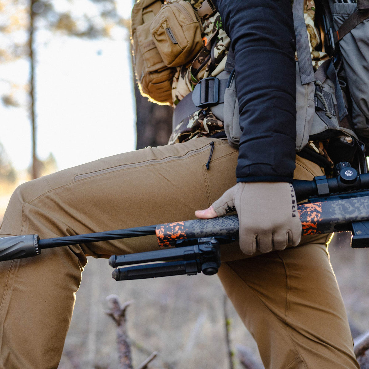 Tan outdoor pants featuring a horizontal zippered thigh pocket, worn by a person holding a camouflaged rifle.