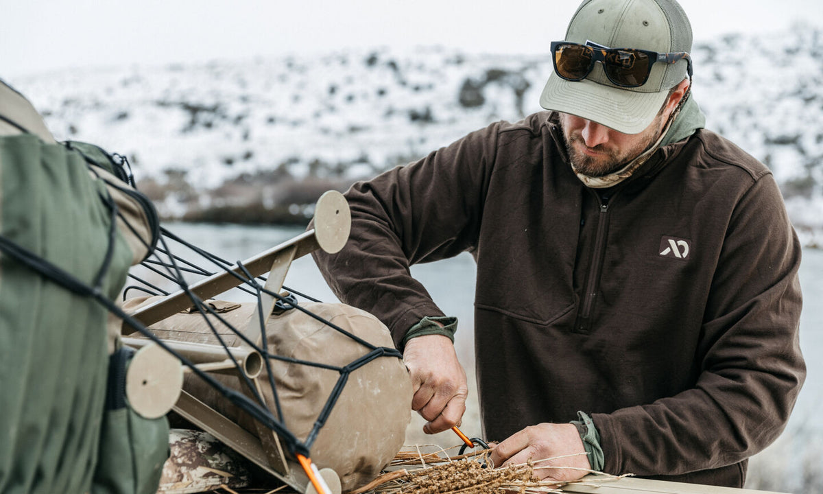 Man wearing brown quilted quarter-zip fleece with white logo, adjusting outdoor gear.