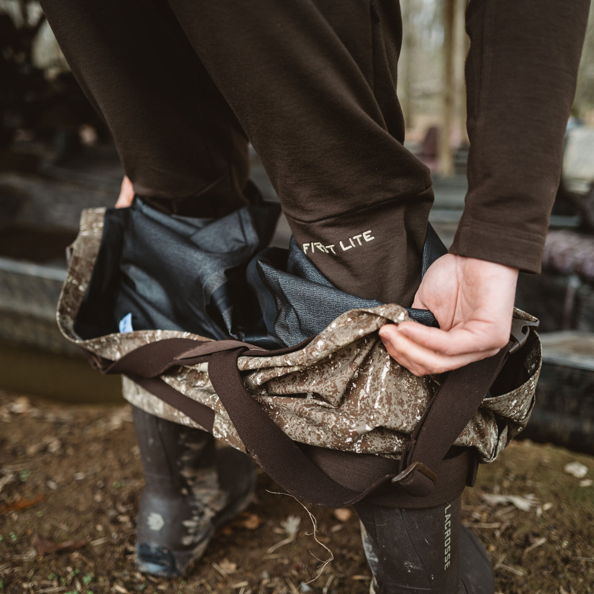 Person adjusting camouflage waders with dark brown straps over brown First Lite pants and dark Lacrosse boots.