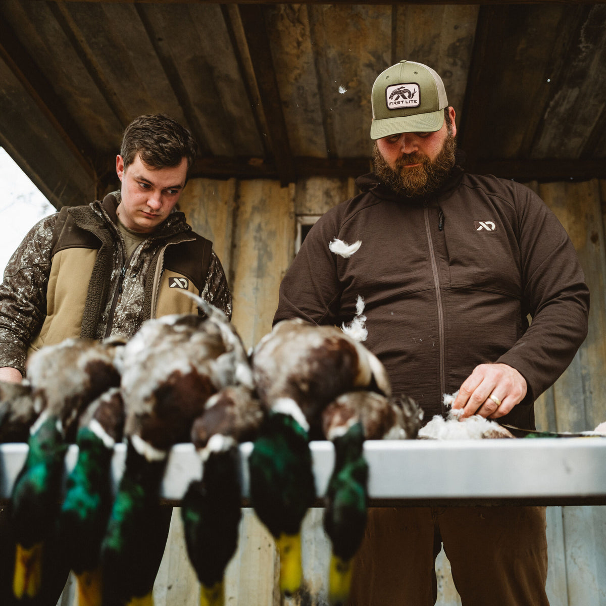 A man in a dark brown First Lite zip-up jacket and olive green First Lite baseball cap plucks feathers from ducks, beside a man in a tan First Lite vest.