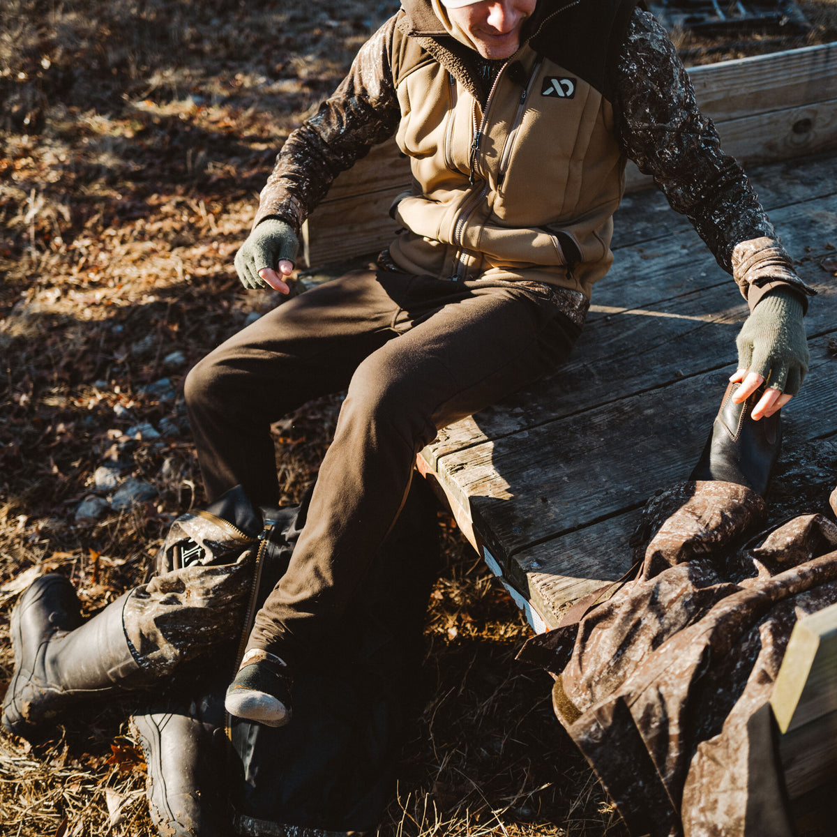 A person adjusts a tall, dark brown outdoor boot worn over camouflage pants.