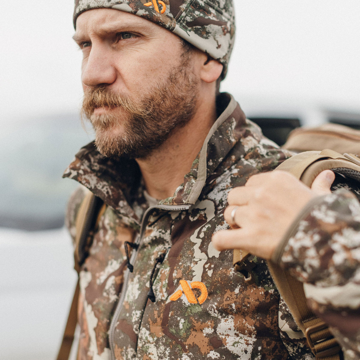 A man wears a brown, green, and white camouflage jacket with an orange logo and a matching beanie.