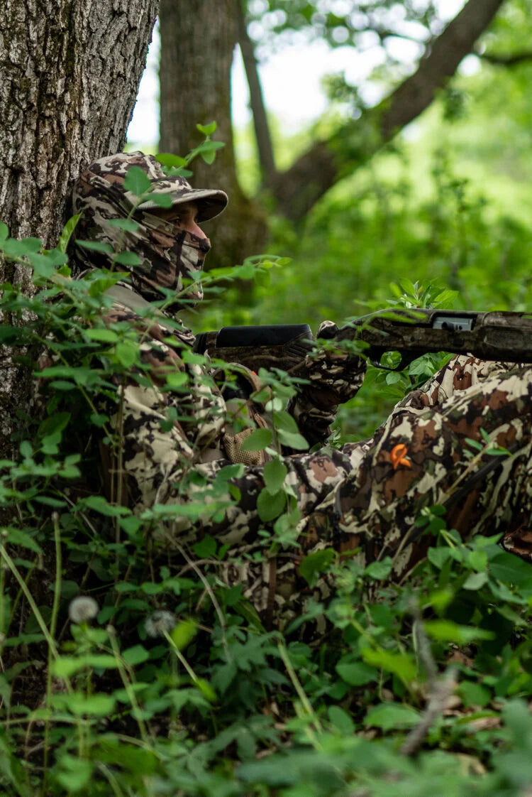 Camouflaged person in mask and cap, holding rifle, concealed in forest foliage.