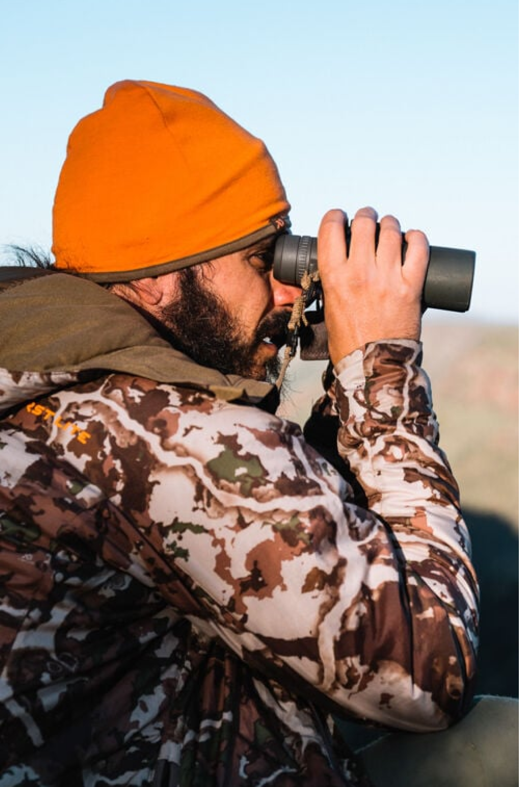 Bearded man in orange beanie & First Lite camo jacket looking through dark green binoculars.