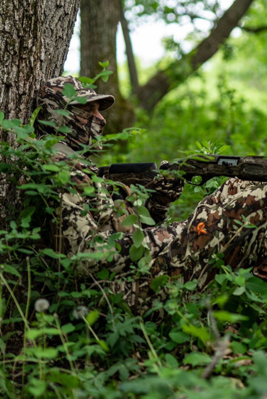 Hunter in brown/tan patterned camo outfit, face mask, holding rifle, leaning on tree.