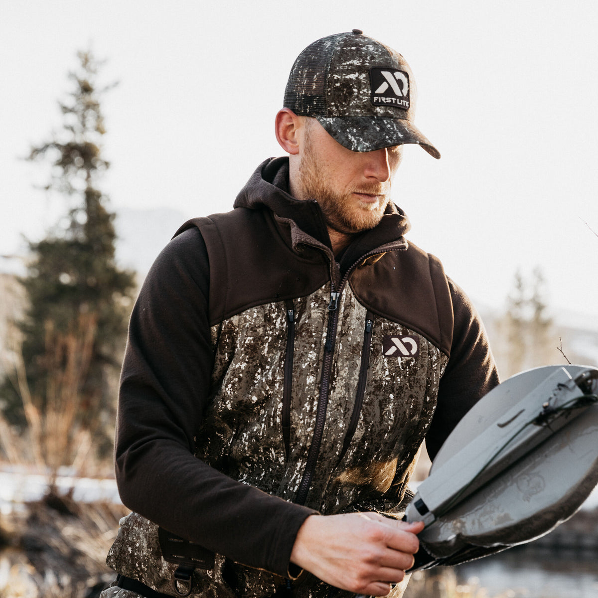 A man wears a First Lite speckled camouflage hunting vest with dark brown shoulders and a matching trucker hat, holding a grey folding object.