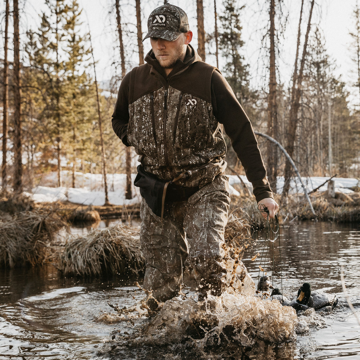 A man in a brown hooded hunting jacket with a camouflage torso and matching camouflage pants wades through muddy water, holding duck decoys.