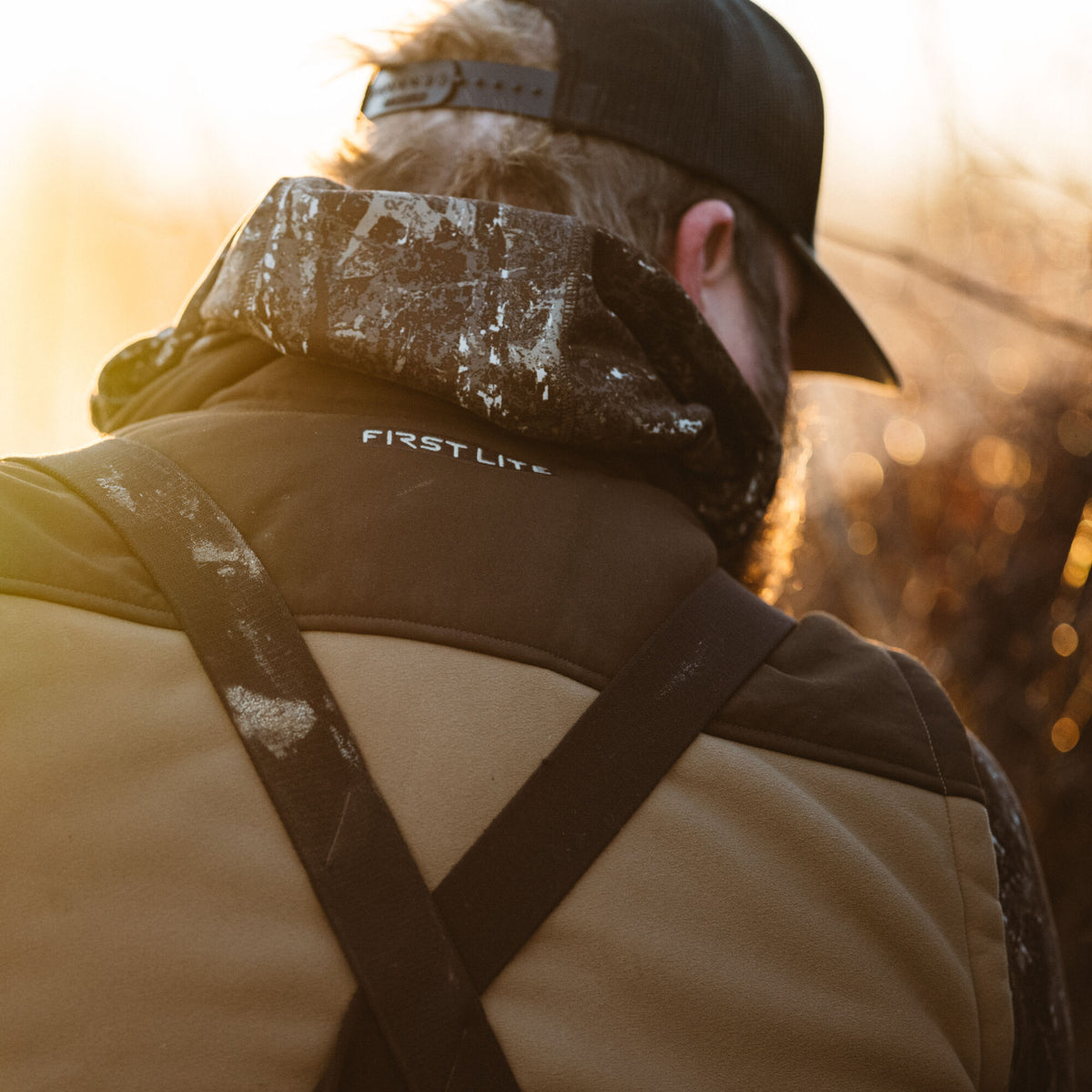 Back of a person wearing a First Lite dark jacket with a camouflage hood and tan bibs with dark shoulder straps.