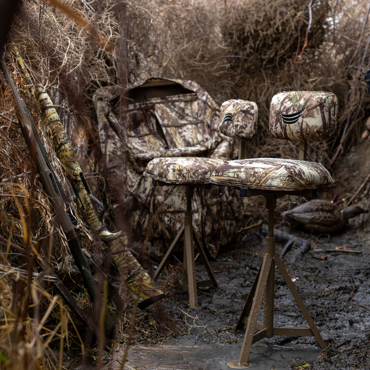 Two camouflage swivel hunting chairs with black logos and a matching portable blind are set up with two rifles on muddy ground.