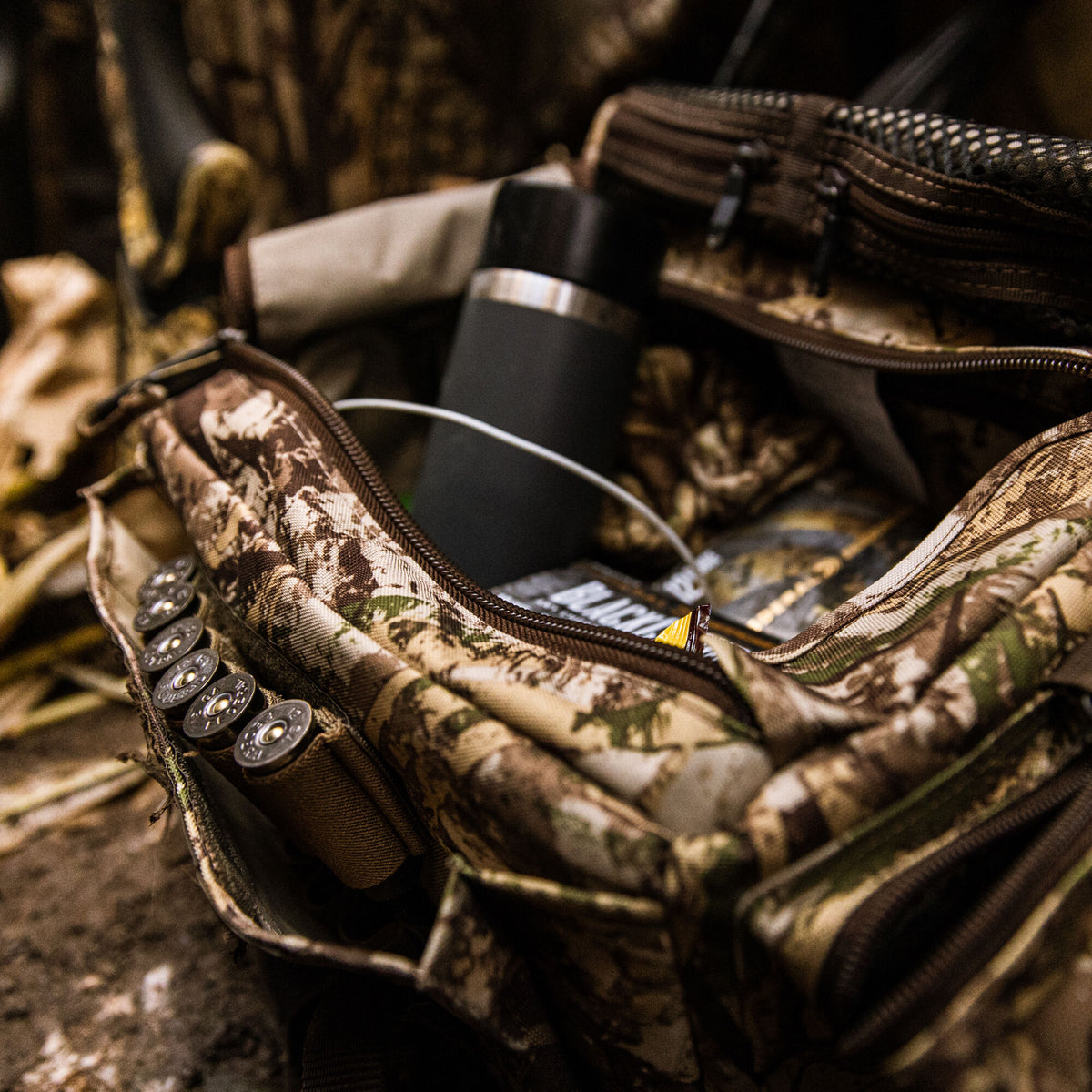 A camouflage gear pouch lies open, revealing shotgun shells in elastic loops, a dark gray thermos, and other supplies inside.