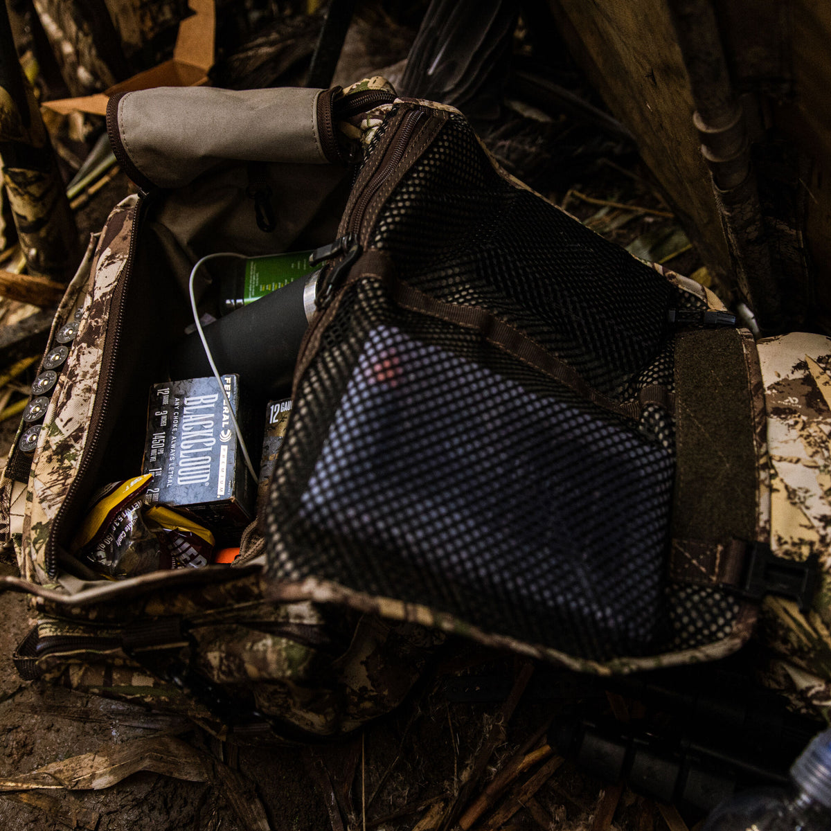 Camouflage gear bag with an open main compartment showing shotgun shells, candy, and a black bottle, plus a mesh pocket and external shell loops.