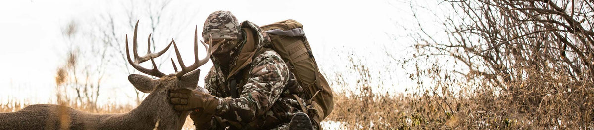 Camo hunting jacket, hat, gloves, and tan backpack on hunter holding a buck.