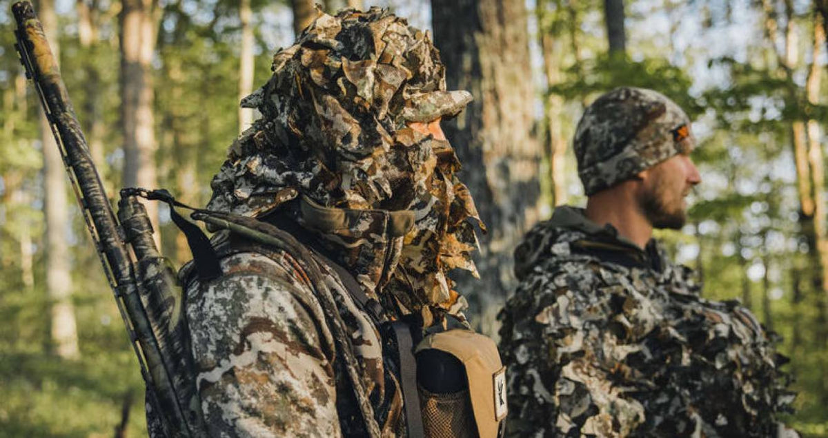 Hunter in a green/brown leafy ghillie suit with a camouflaged rifle on their back.