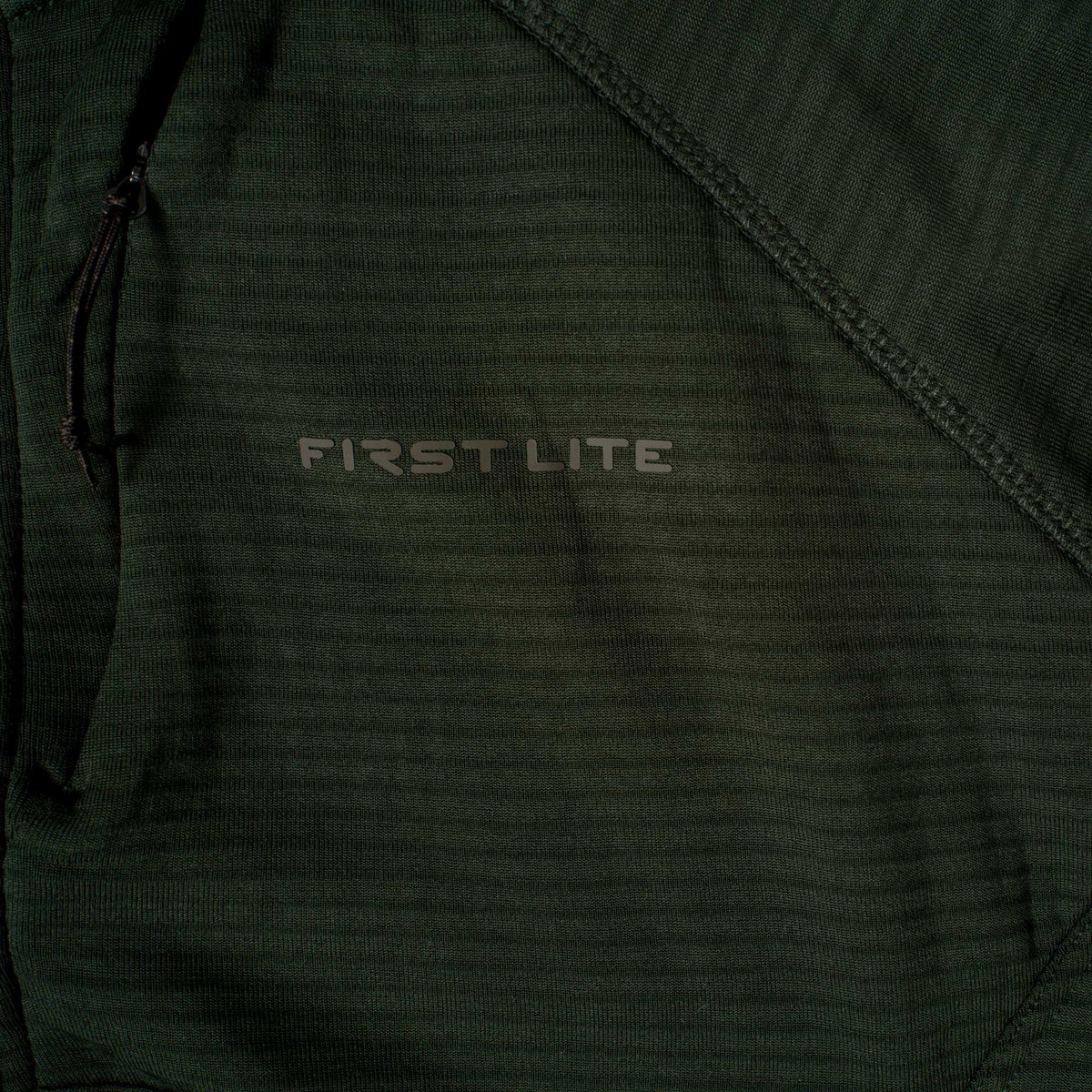 Deep forest green textured long-sleeved top featuring a 'FIRST LITE' logo, a zippered chest pocket, and diagonal seam detail.