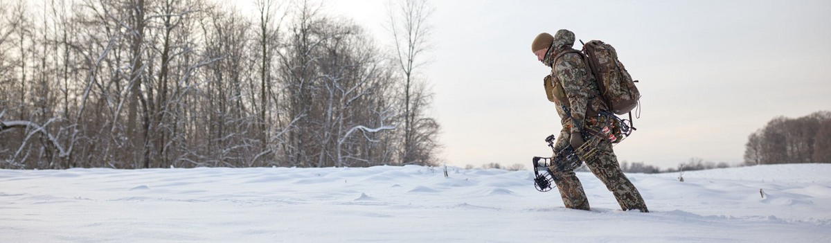 Camo hunter with matching backpack and bow walks across snowy field.