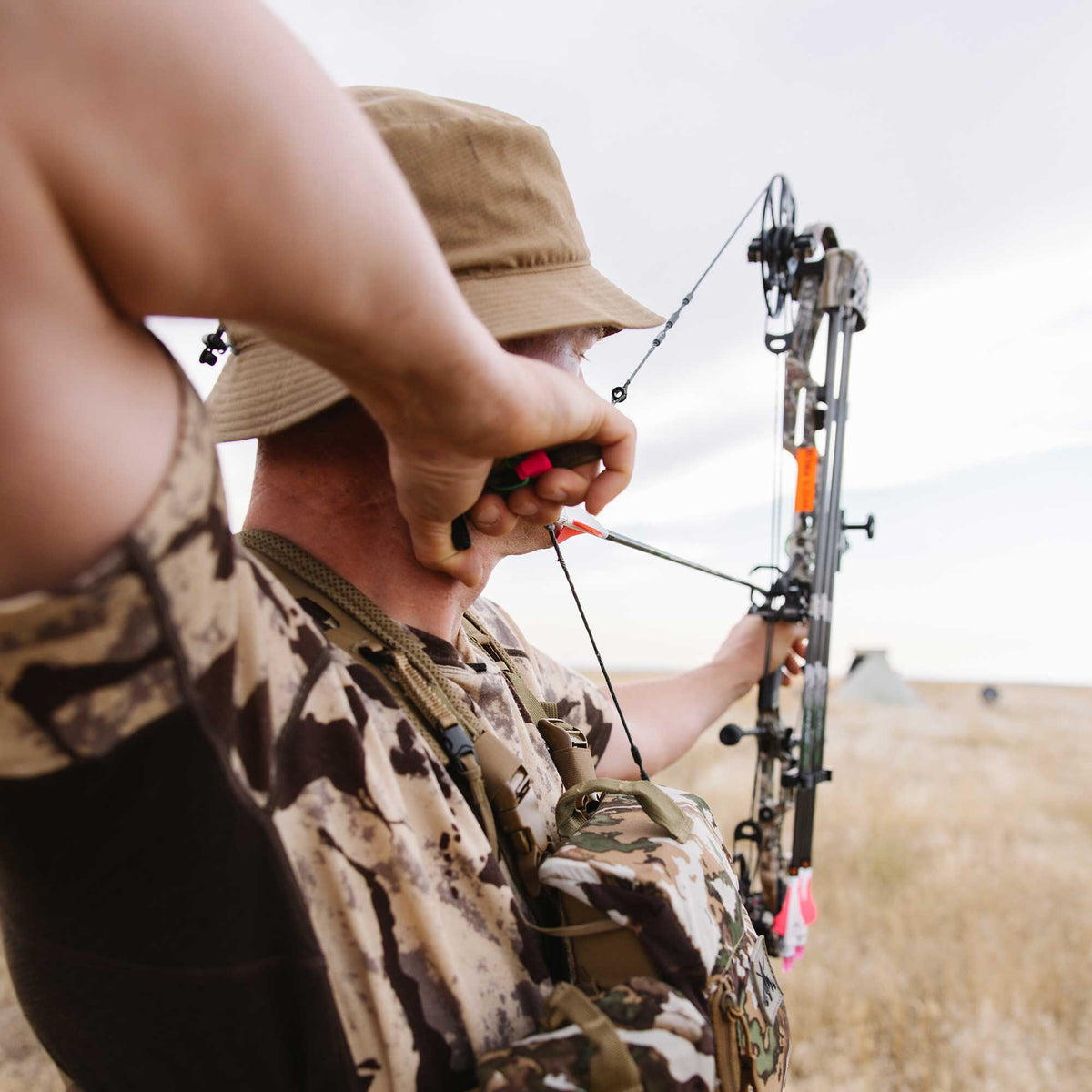 A person in a tan hat and camouflage vest aims a drawn camouflage compound bow with an orange-fletched arrow.