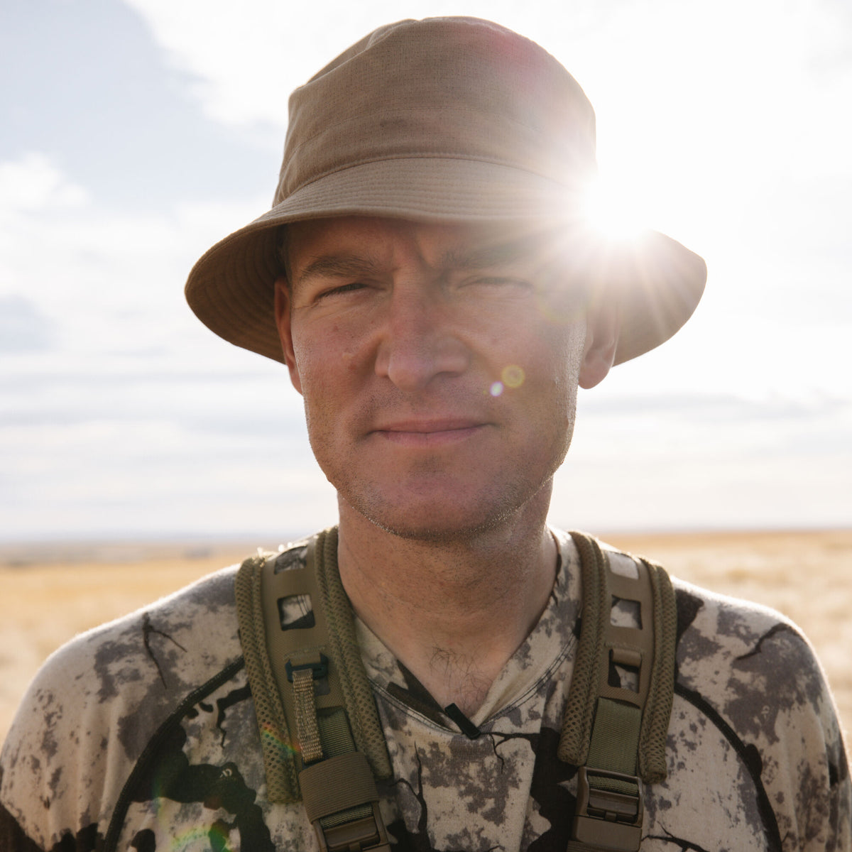 Man wears a tan fabric bucket hat, a grey and black camouflage long-sleeve shirt, and olive green mesh backpack straps.