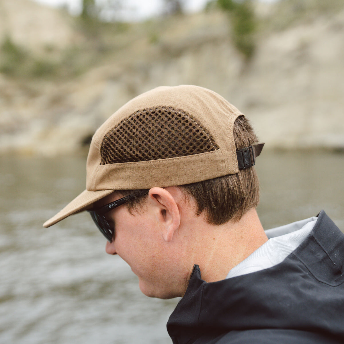 A tan 5-panel cap featuring dark mesh ventilation panels and a black adjustable strap, worn by a person.