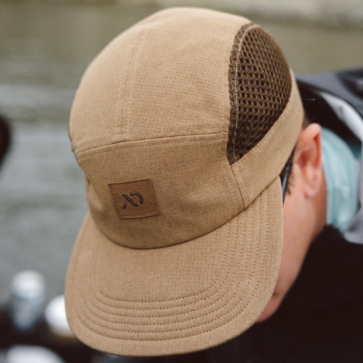 Tan five-panel cap with perforated fabric, dark brown mesh side panels, a stitched brim, and a square logo patch.