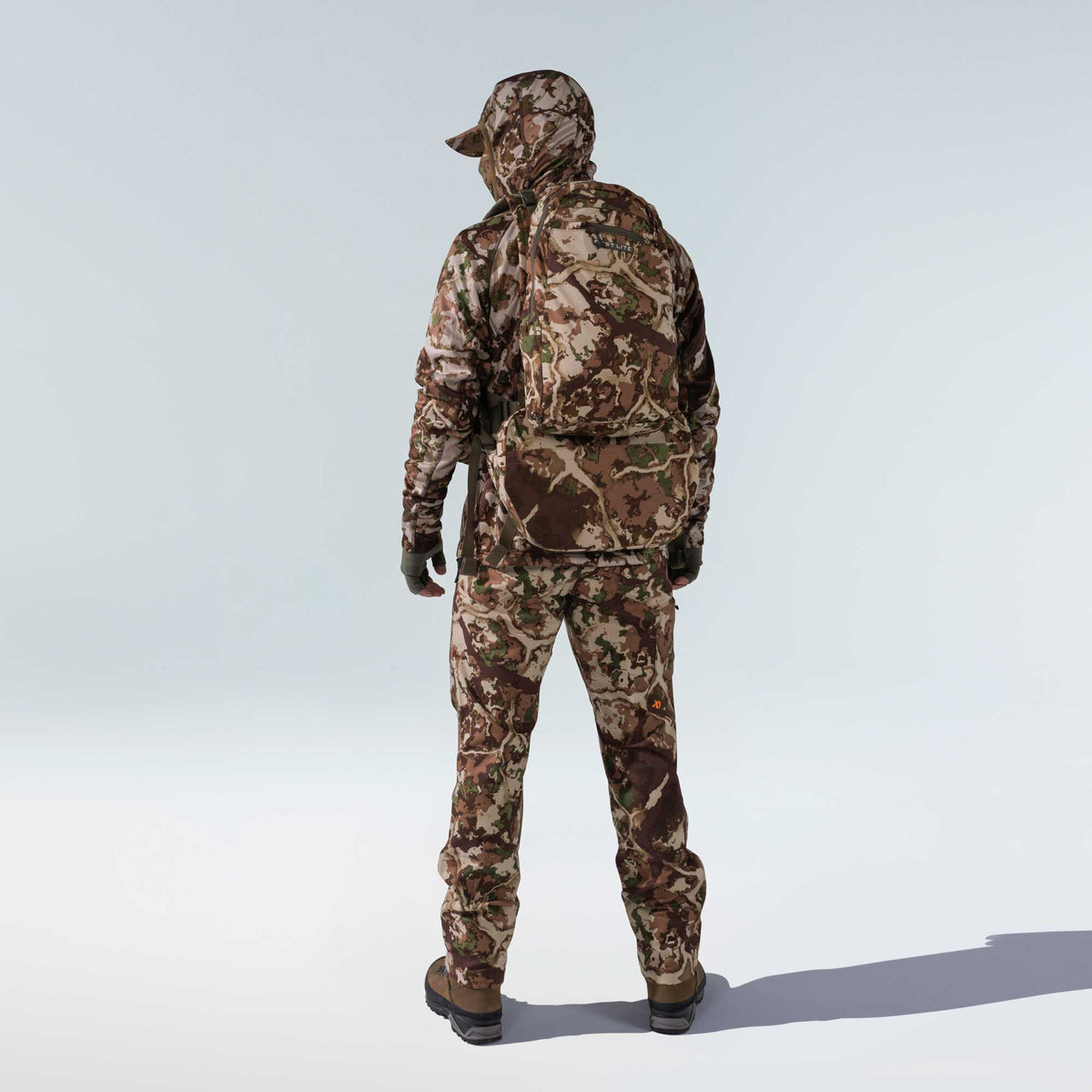 Rear view of a person wearing a First Lite brown, tan, and green camouflage hunting suit, including a hooded jacket, pants, cap, gloves, matching backpack, and brown boots.