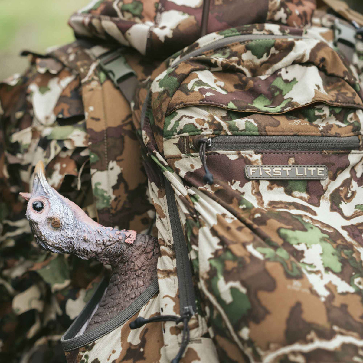 A First Lite camouflage hunting backpack with a turkey decoy's head peeking from a side pocket.