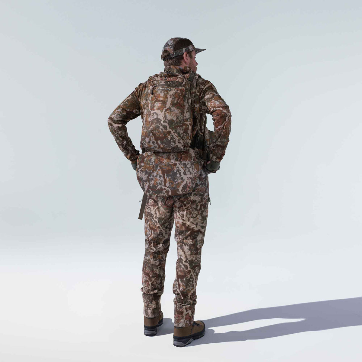 A man wears a full First Lite camouflage hunting outfit, featuring a cap, backpack, and waist pack, standing with his back to the viewer in a brown, green, and tan pattern.