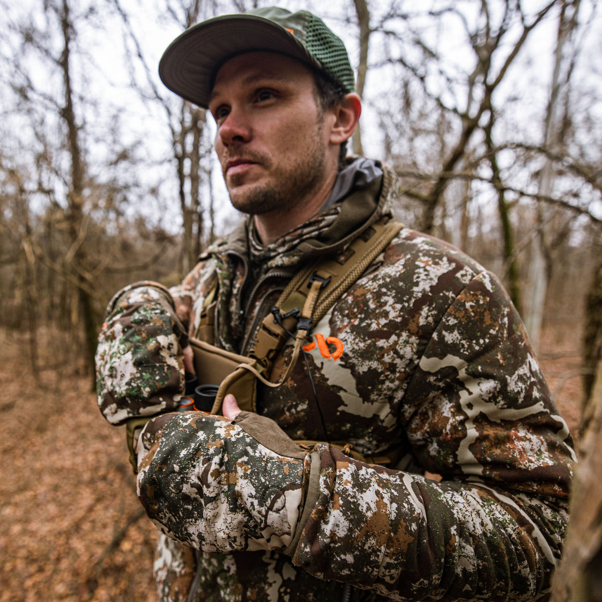Man in a white, brown, and green camouflage hunting jacket with an orange logo on a tan chest rig and matching gloves.