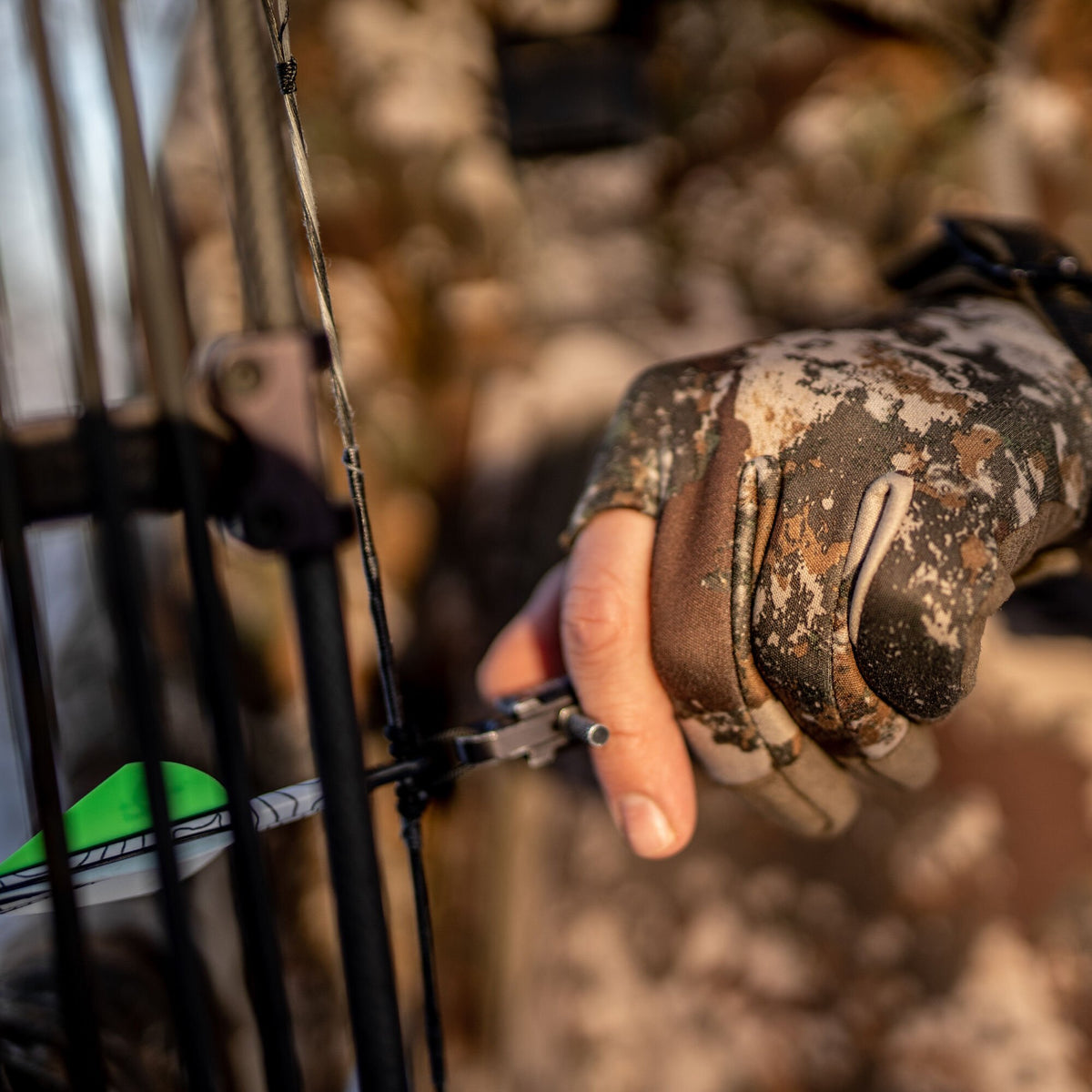 Camouflage hunting glove with an exposed fingertip gripping a bow release aid, with a neon green fletched arrow in the foreground.