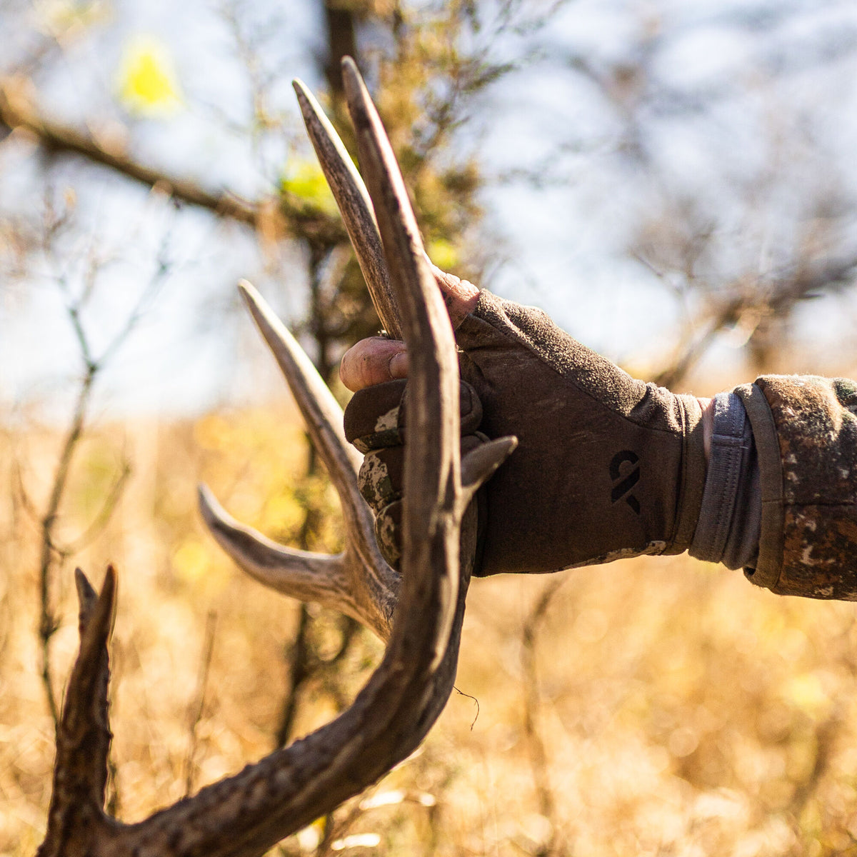Brown outdoor glove with a black logo and grey camo cuff, holding deer antlers.
