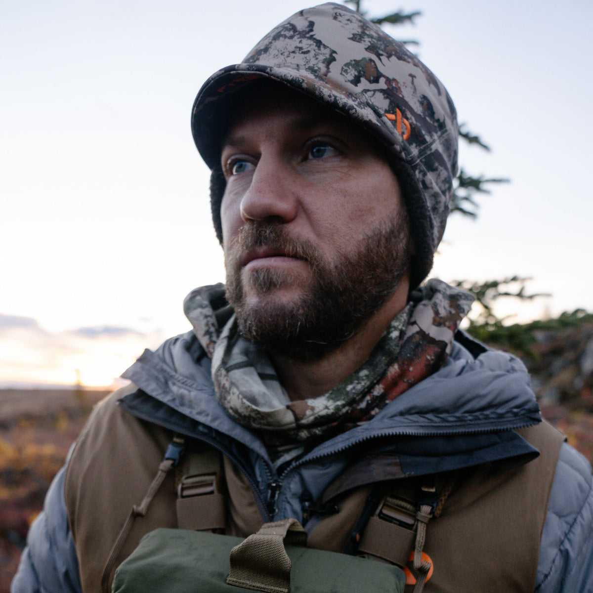 Camouflage winter hat featuring a brim, dark ear flaps, and an orange brand logo.