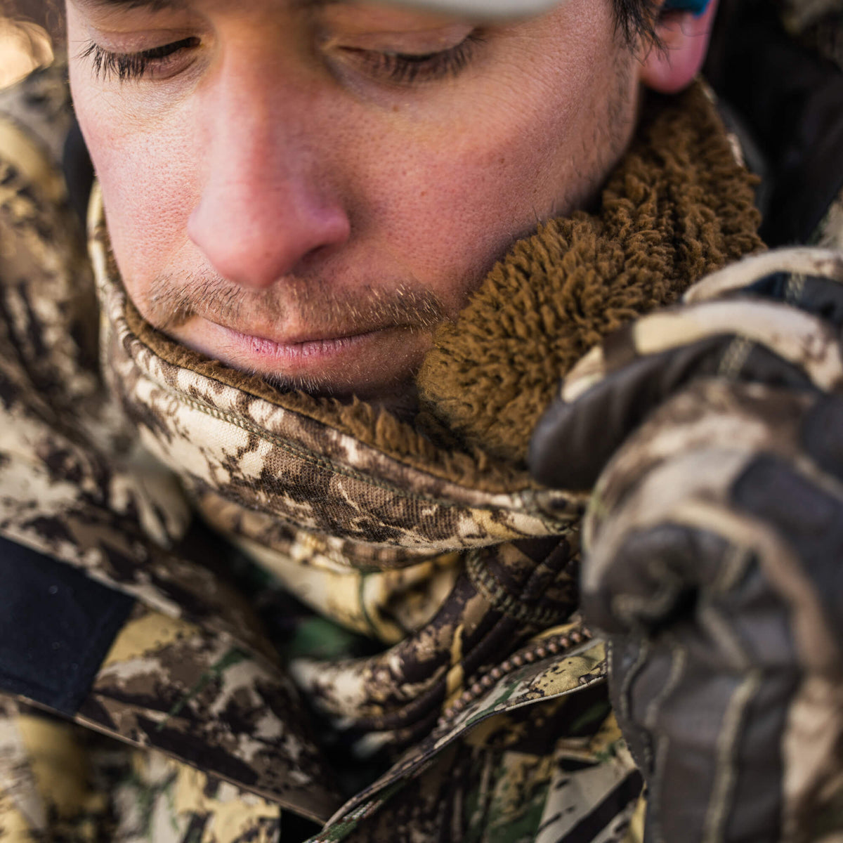 Close-up of a person wearing an earth-toned camouflage jacket with a brown sherpa-lined collar, being adjusted by a gloved hand.