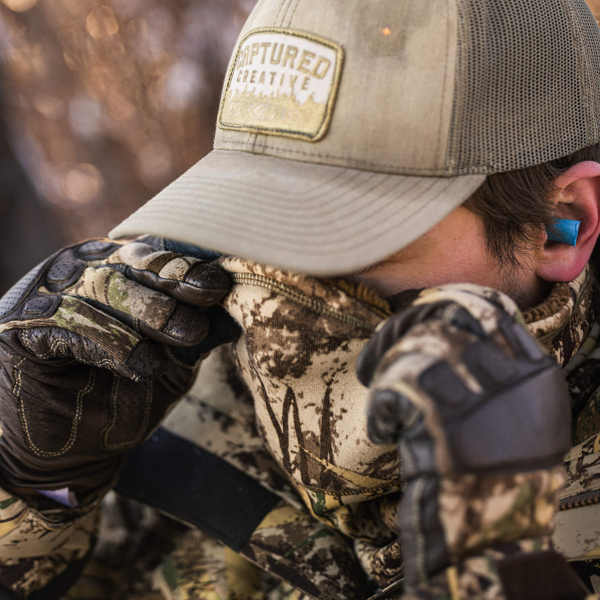 A person wears a tan trucker hat and camouflage gloves, with a bright blue earplug visible in their right ear.