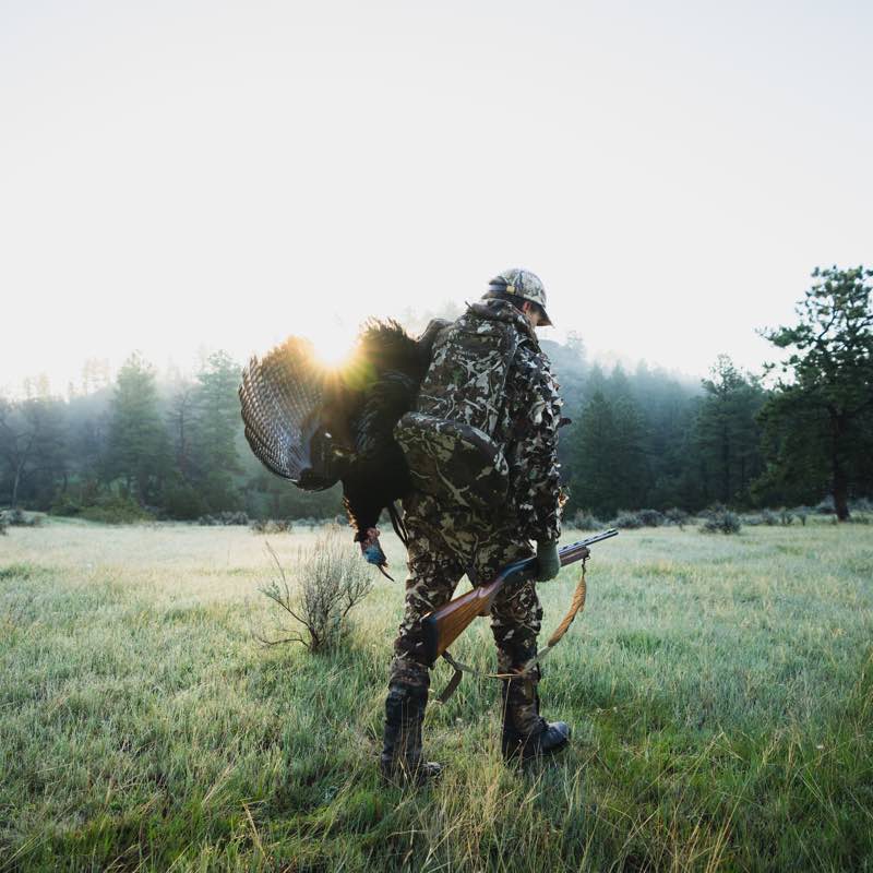 Hunter in camouflage apparel carrying shotgun and turkey in a misty field at sunrise.