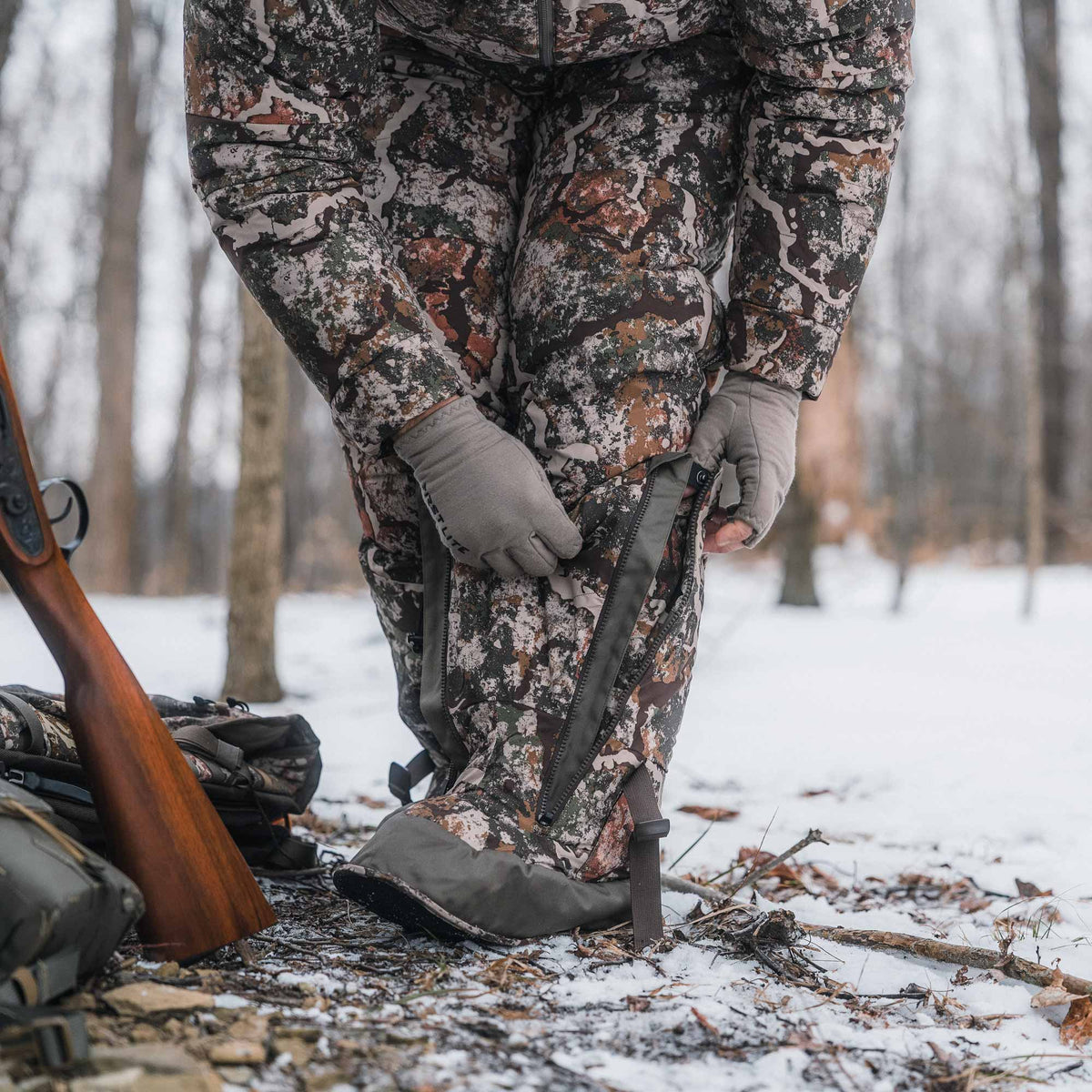 A person wearing a camouflage hunting jacket and pants with gray gloves is standing in a snowy forest, adjusting a black strap on the pants. A rifle and backpack are visible on the ground nearby.