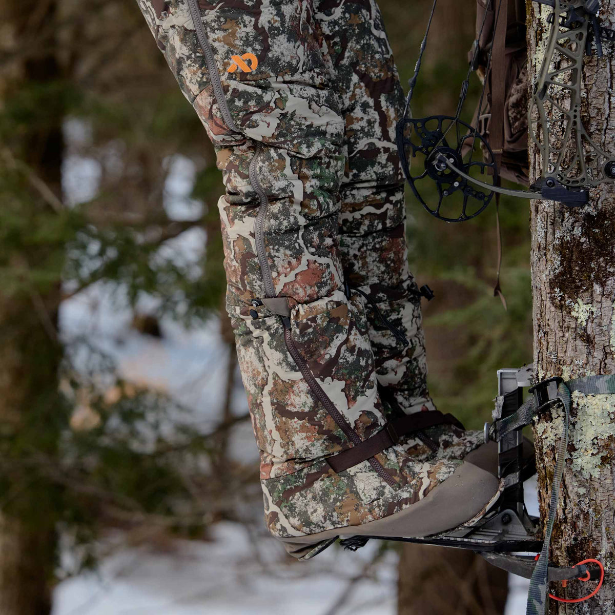 A close-up of a person wearing camouflage hunting pants and a boot, suspended in a tree stand with a bow and compound bow visible in the background of a snowy forest.
