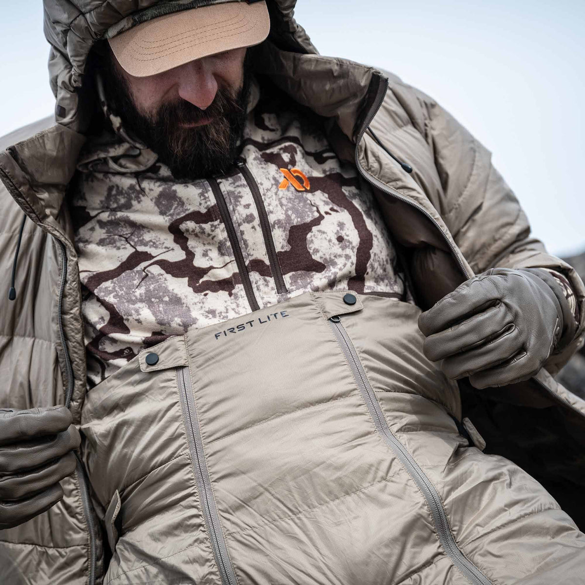 A person wears coyote tan First Lite puffy insulated bibs with full-length side zippers and a matching hooded jacket over a camouflage shirt.