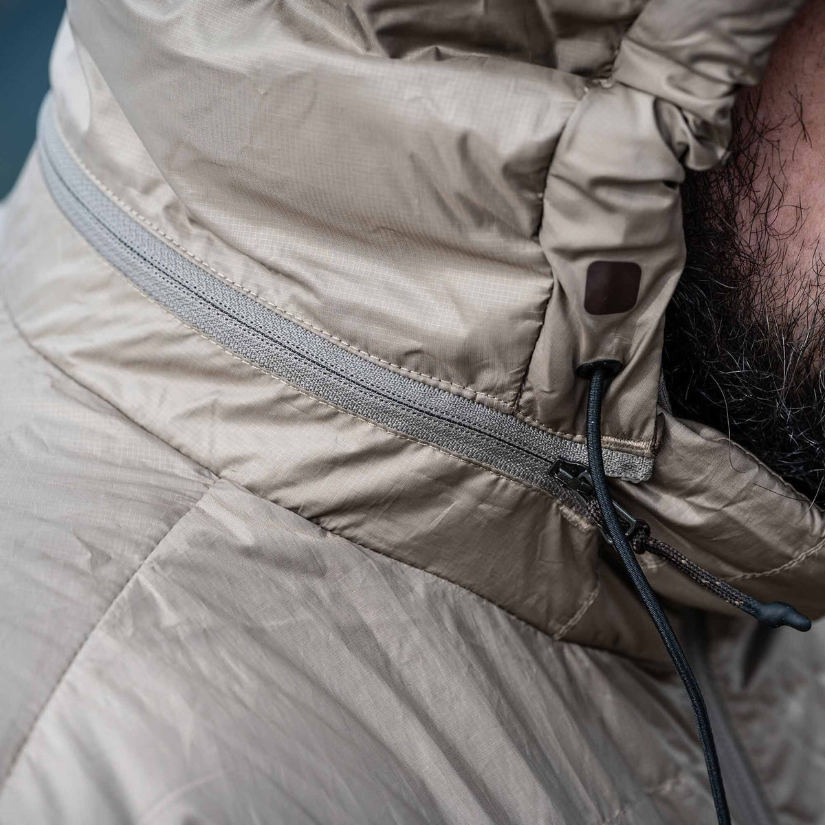 Light khaki ripstop hooded jacket with a grey zipper, hood drawstring, and a dark square patch.