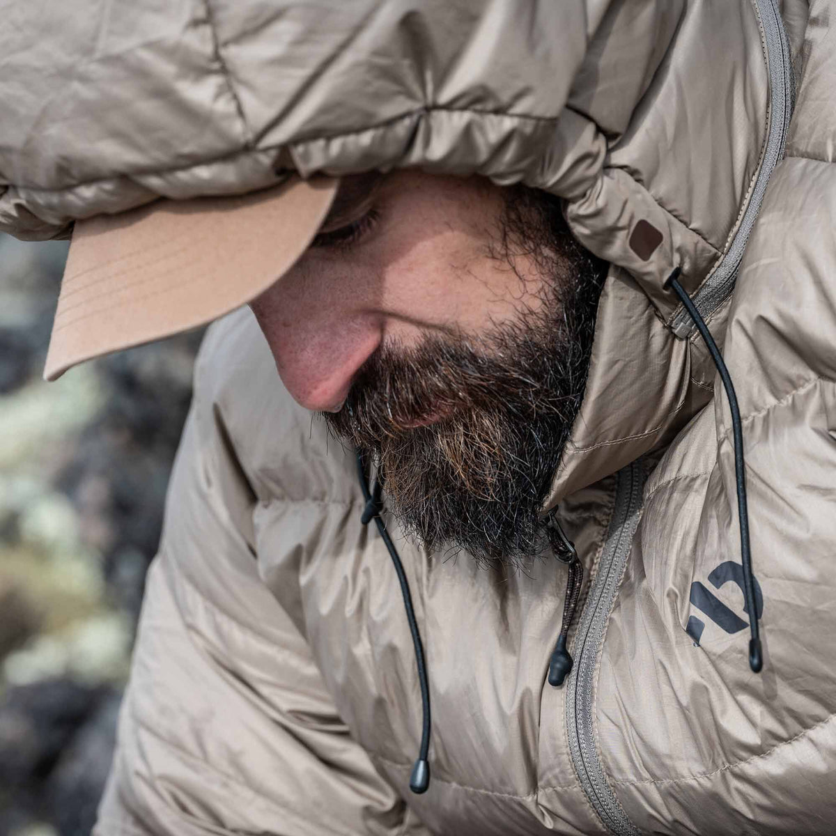 Tan hooded puffer jacket on a person, featuring a front zipper, drawstrings, and a dark logo on the right chest.