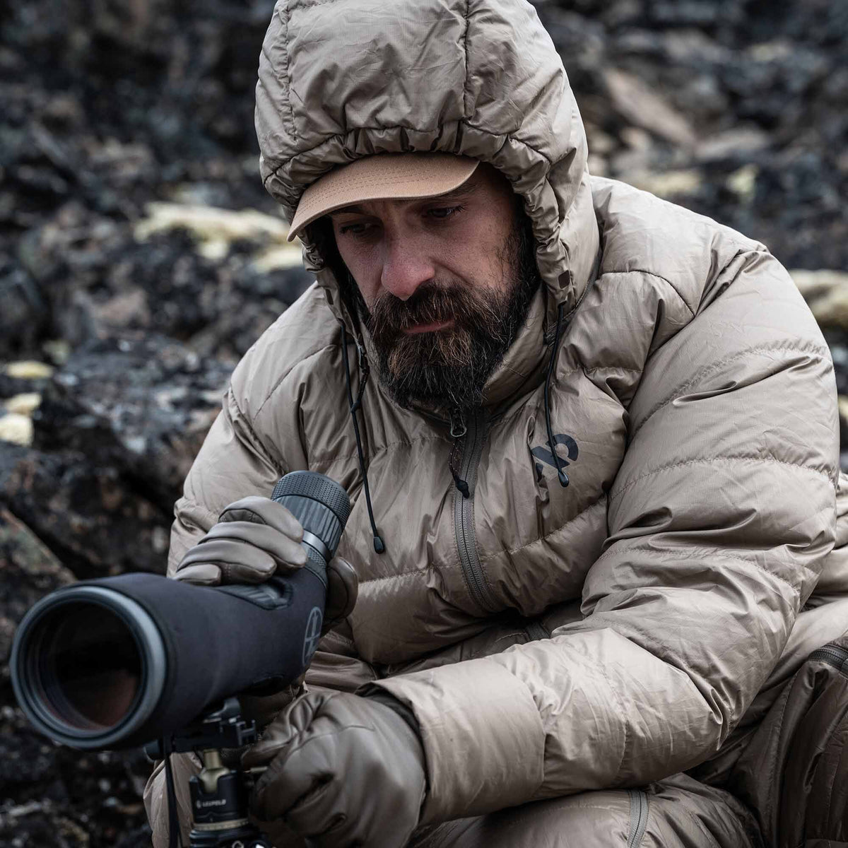 Tan quilted hooded jacket with a black chest logo, worn by a man looking through a spotting scope.