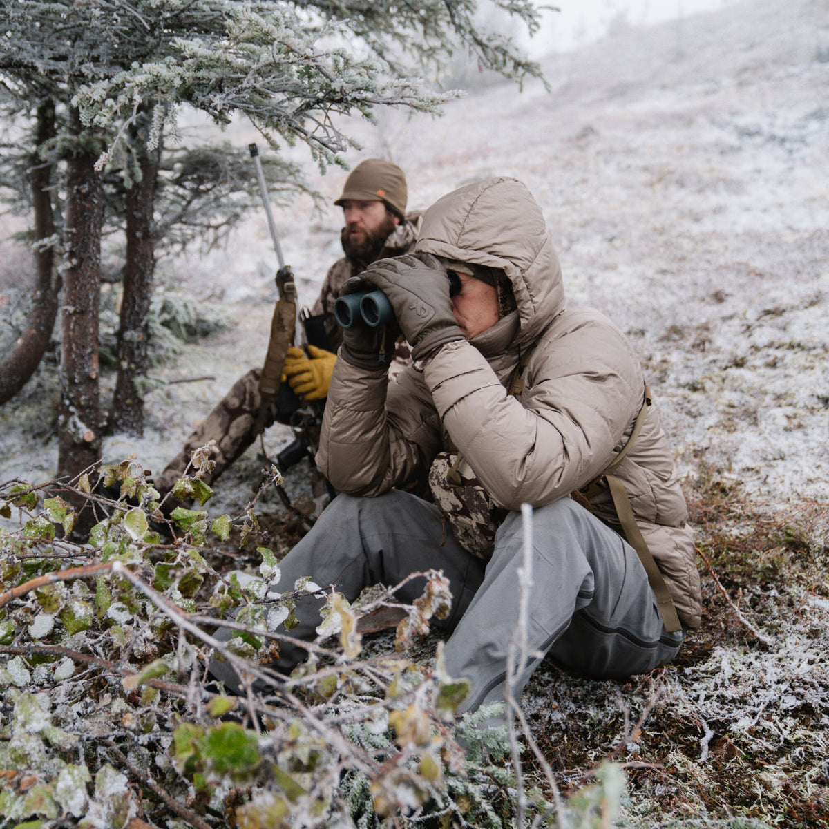 Fawn hooded puffer jacket with a slight sheen, worn by a person looking through binoculars in a snowy landscape.