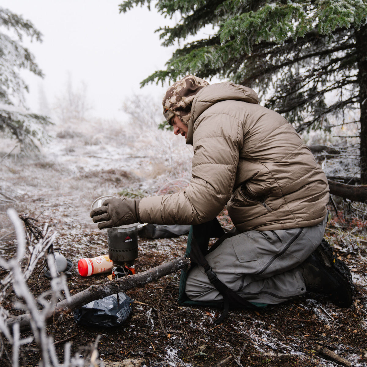 Tan puffer jacket with horizontal baffles worn by a person cooking in a snowy forest.