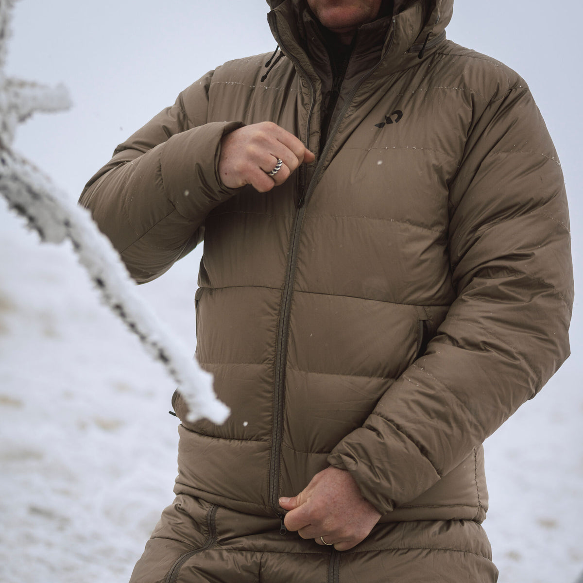 A person zipping a brown hooded puffer jacket with a black chest logo in a snowy environment.