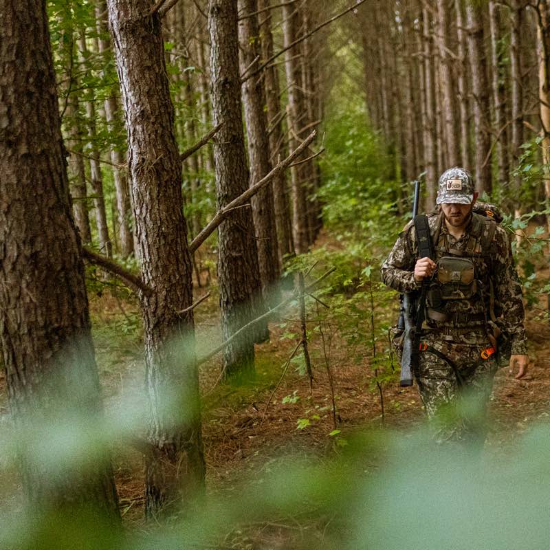 Brown/green camouflage hunting uniform with chest rig and hat worn by a man with a rifle.