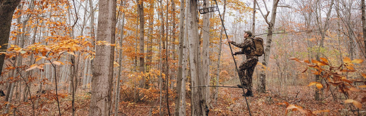 Camo hunter uses dark tree climbing sticks to ascend a tree with a tan backpack.