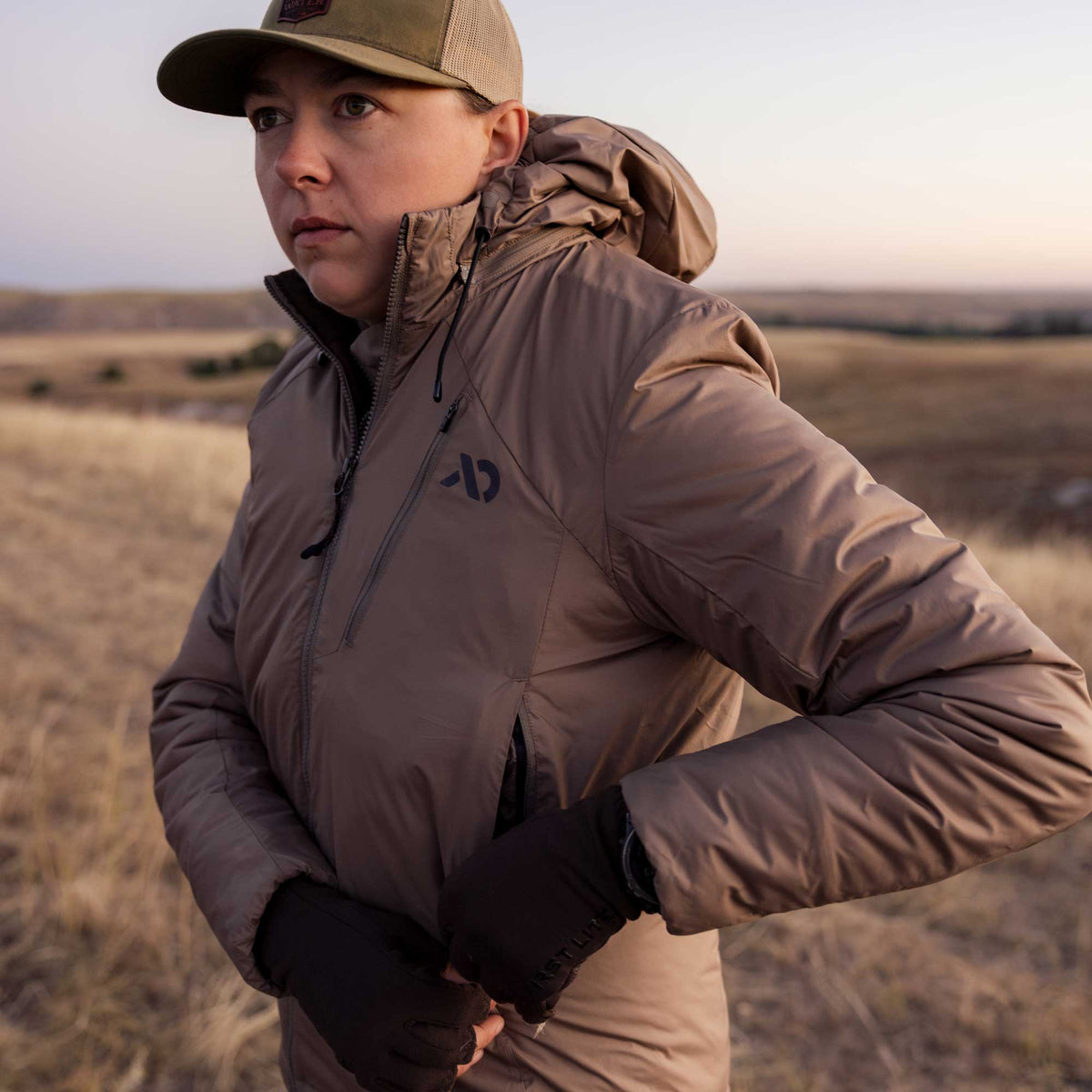 A woman wears a tan hooded insulated jacket with a chest logo, front zipper, and zippered hand pockets.
