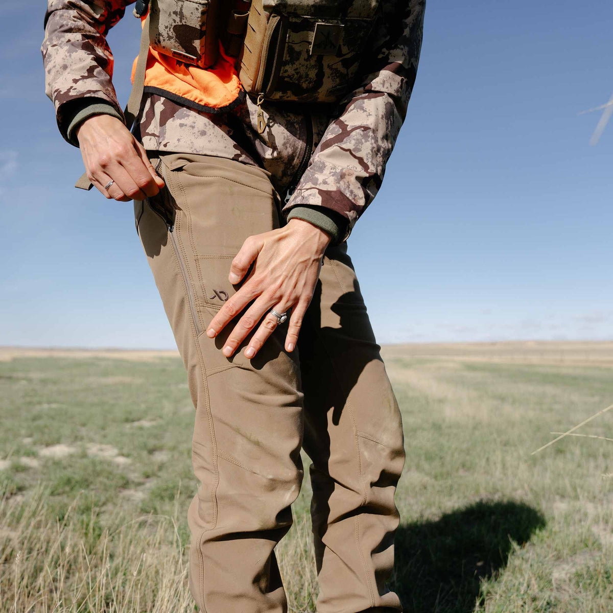 Tan outdoor pants with zippered side pockets and a thigh logo, being unzipped by a person in a sunny field.